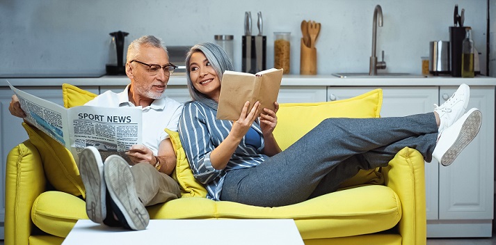 cheerful Asian woman showing book to elderly husband holding newspaper